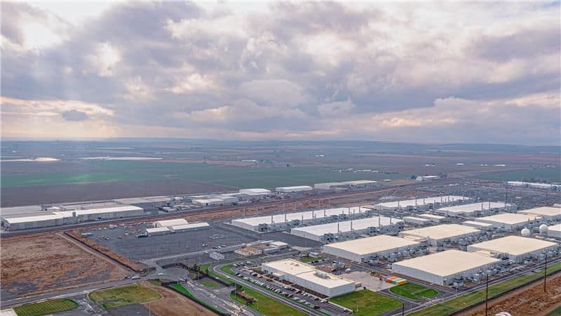 Aerial view of a Microsoft data center campus surrounded by farmland, with rows of low white server buildings, backup generator yards and electrical substations under a cloudy sky.
